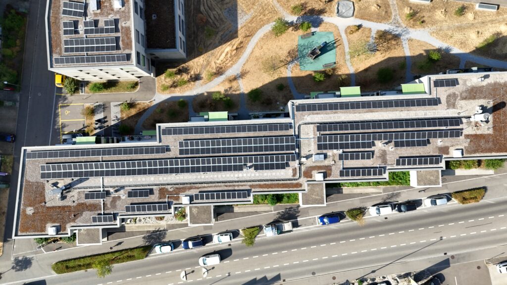Aerial view of a building complex with many rooftop solar panels, surrounding paths, and a central green playground area from above.