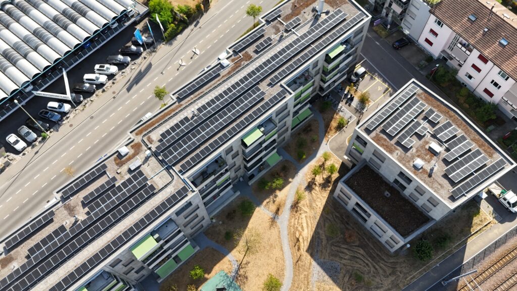 Aerial view of multi-story buildings with extensive rooftop solar panels and a street lined with parked cars nearby.