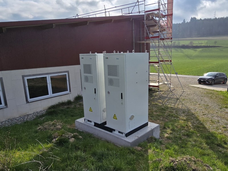 Two gray electrical cabinets on a concrete pad outside a building, with scaffolding nearby and a green field in the background.