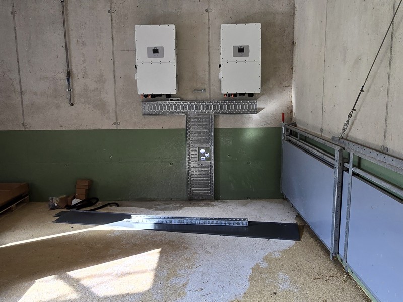 Industrial utility area with two white electrical cabinets on a concrete wall, metal cable tray forming a T shape, and a sunlit floor.
