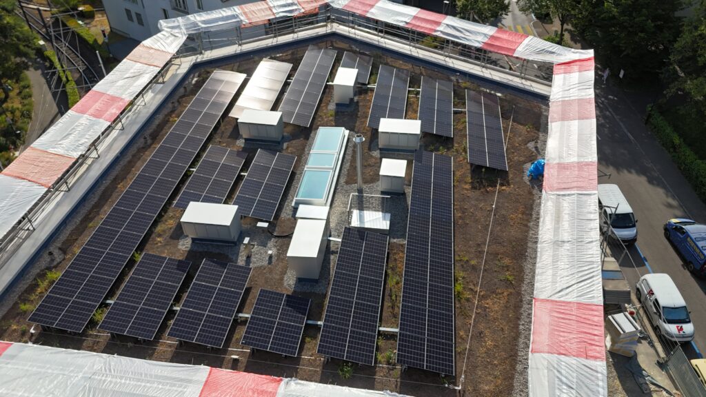 A rooftop covered with many dark solar panels arranged in rows, surrounded by white and red striped tarps and nearby parked cars on the street below.