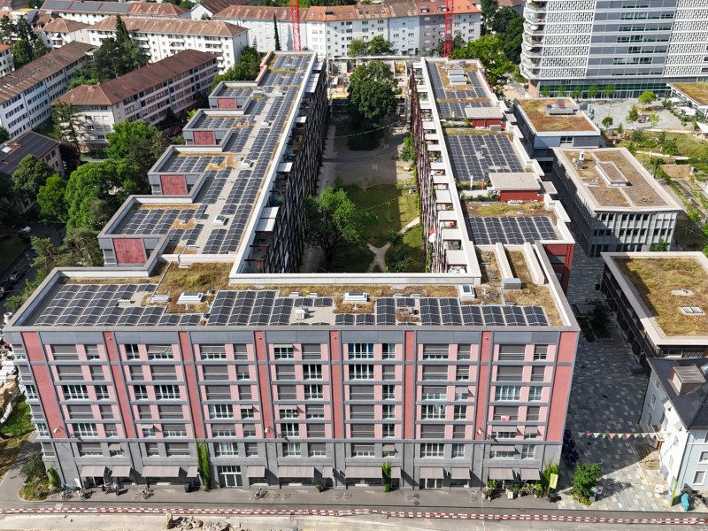 U-shaped residential complex with pink and gray facade, solar panels on flat roofs, and a green central courtyard seen from above, in a city block with nearby buildings and cranes nearby.