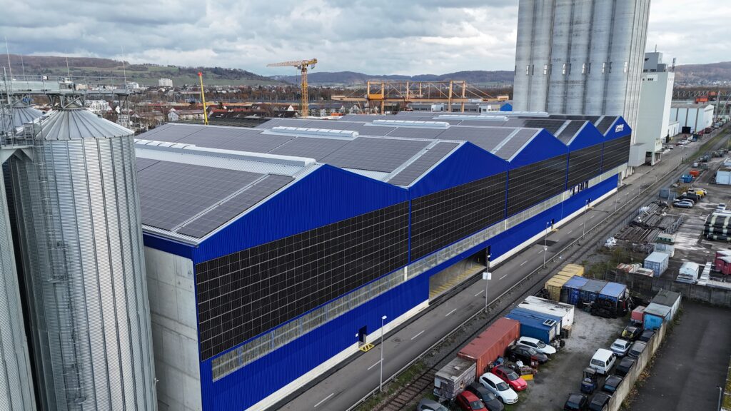 Aerial view of a blue and black industrial warehouse with solar panels on the roof and tall silos on the left, plus a road with parked cars nearby.