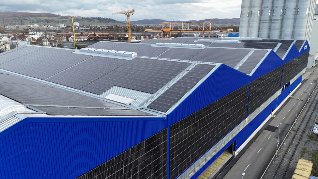 Wide view of a blue industrial building covered with solar panels and adjacent railway tracks in an urban area with hills in the background.