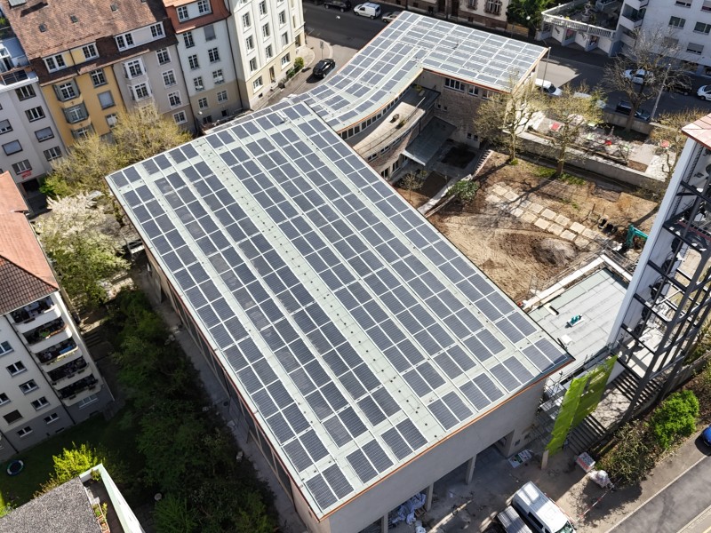 Aerial view of a large building roof covered with solar panels in an urban area, surrounded by streets and other buildings.