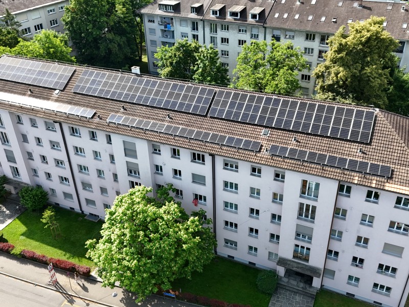 Aerial view of a white apartment building with solar panels lining the brown-tiled roof, surrounded by green trees and a manicured lawn.