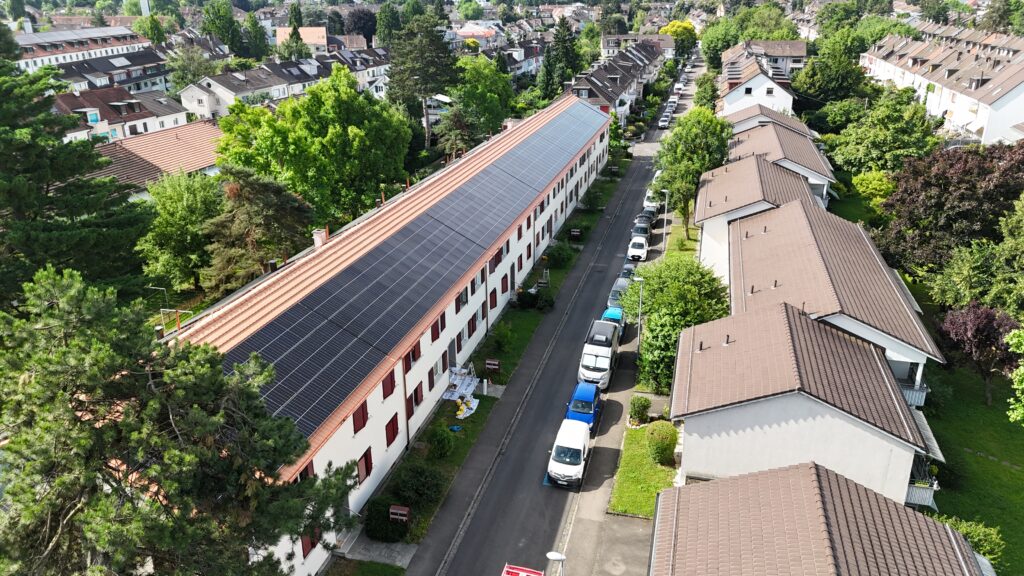 Aerial view of a tree-lined street flanked by long white apartment buildings with red shutters and solar panels on the roof, parked cars along the road.