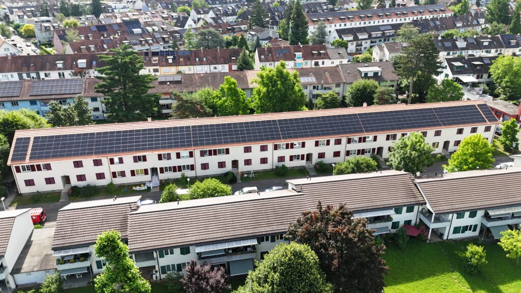 Aerial view of a long white apartment building with a solar-panel roof, surrounded by trees and other houses in a dense neighborhood.