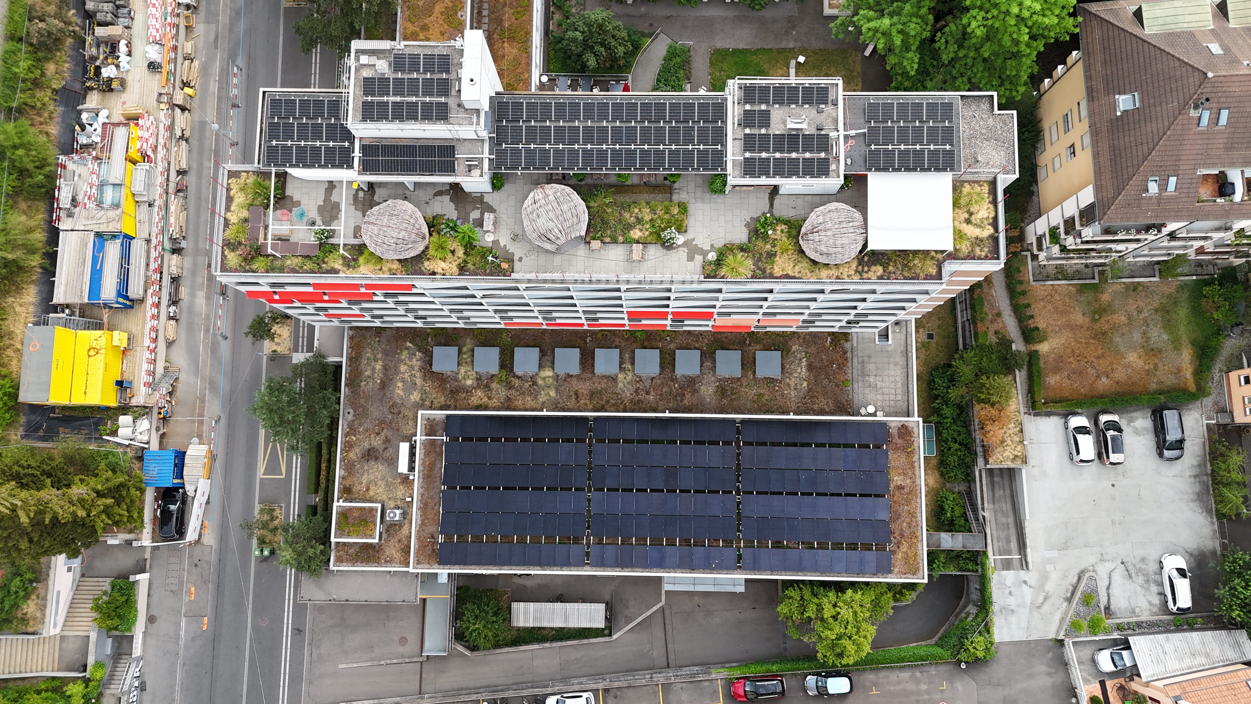 Aerial view of a rooftop building covered with solar panels, courtyard plants, and circular thatched structures between sections; surrounding roads and construction area on left.