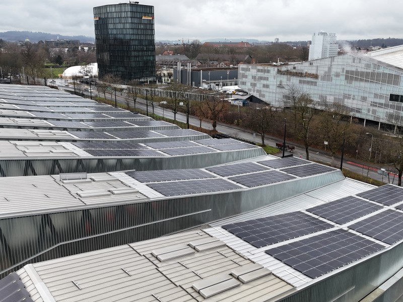 Aerial view of solar panels on sloped metal roofs of an industrial campus, with a cylindrical glass office tower and glass-roofed buildings in the background.