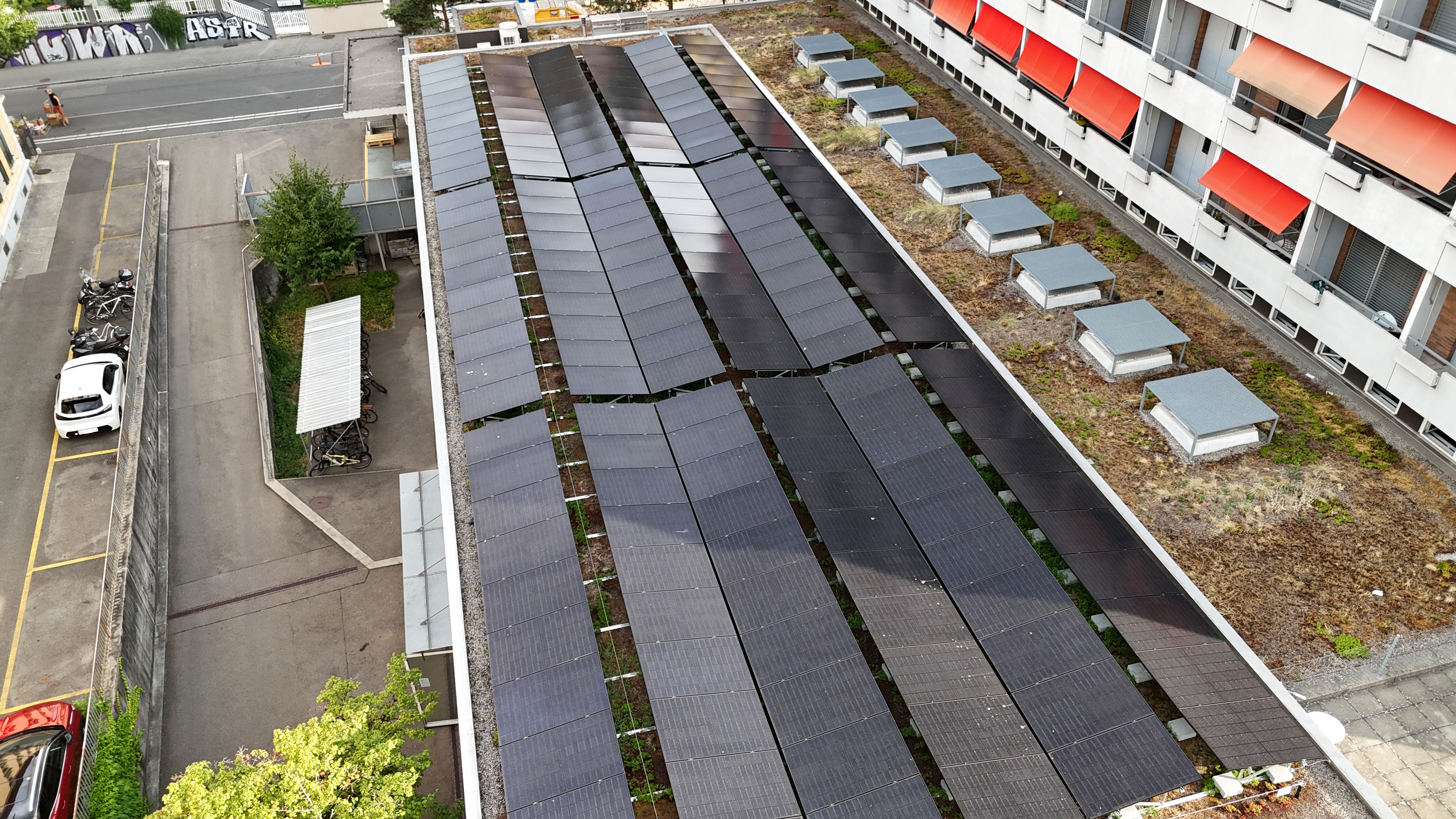 Rooftop solar panels arranged in long rows on a building, with an adjacent courtyard and street below. Aerial view of a building rooftop covered in dark solar panels, flanked by urban streets and balconies with orange awnings.