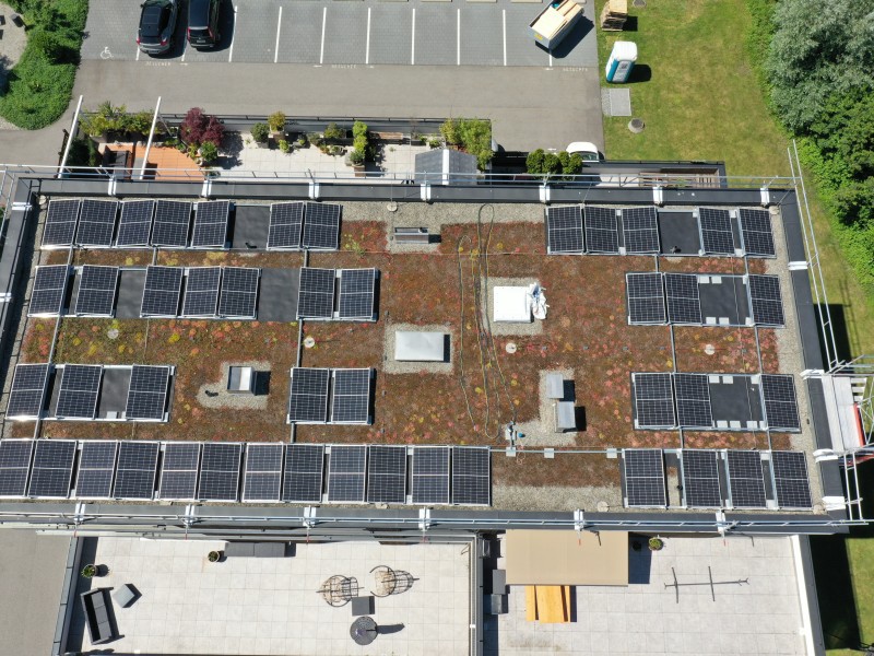 Top-down view of a building roof covered in numerous solar panels arranged in neat rows, with rooftop equipment and surrounding parking lot and greenery nearby.