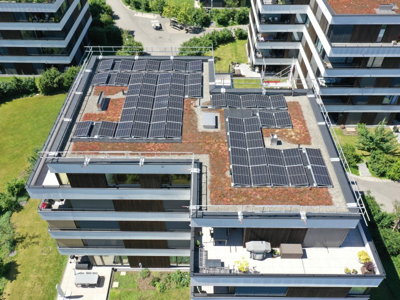 A rooftop covered with numerous solar panels on a modern apartment building, with green lawns and nearby buildings visible from above.