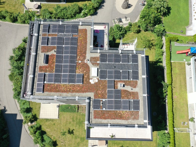 Aerial view of a flat-roof building covered with numerous solar panels arranged in clusters, surrounded by green lawns and trees throughout the campus area.