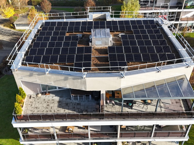 Aerial view of a flat rooftop densely covered with solar panels arranged in rows around a central HVAC unit and metal railing surrounding the edge.