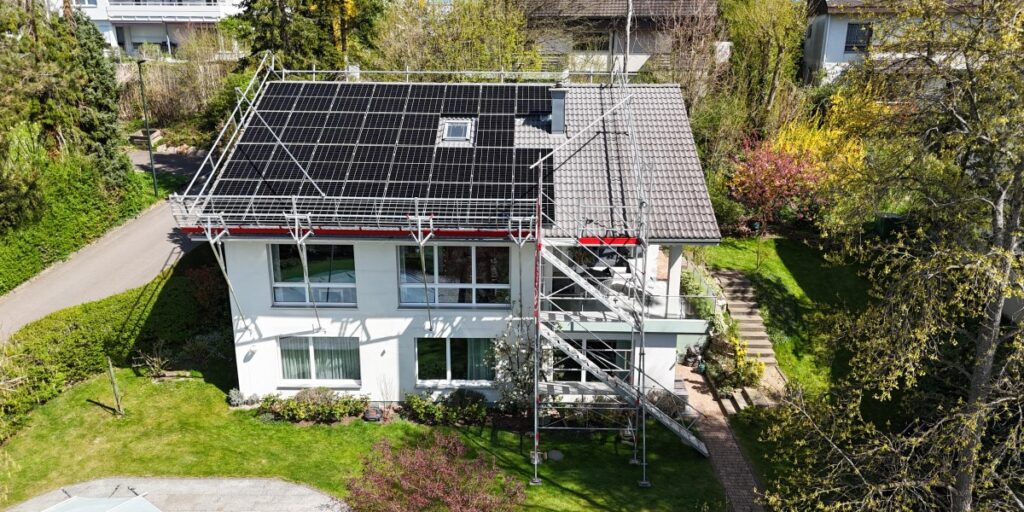 Two-story white house with a large solar panel array on the roof and metal scaffolding around the exterior during renovation.