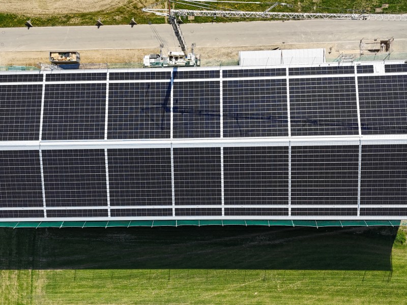 Aerial view of a solar farm with long rows of panels, a crane above, beside a road and a canal.