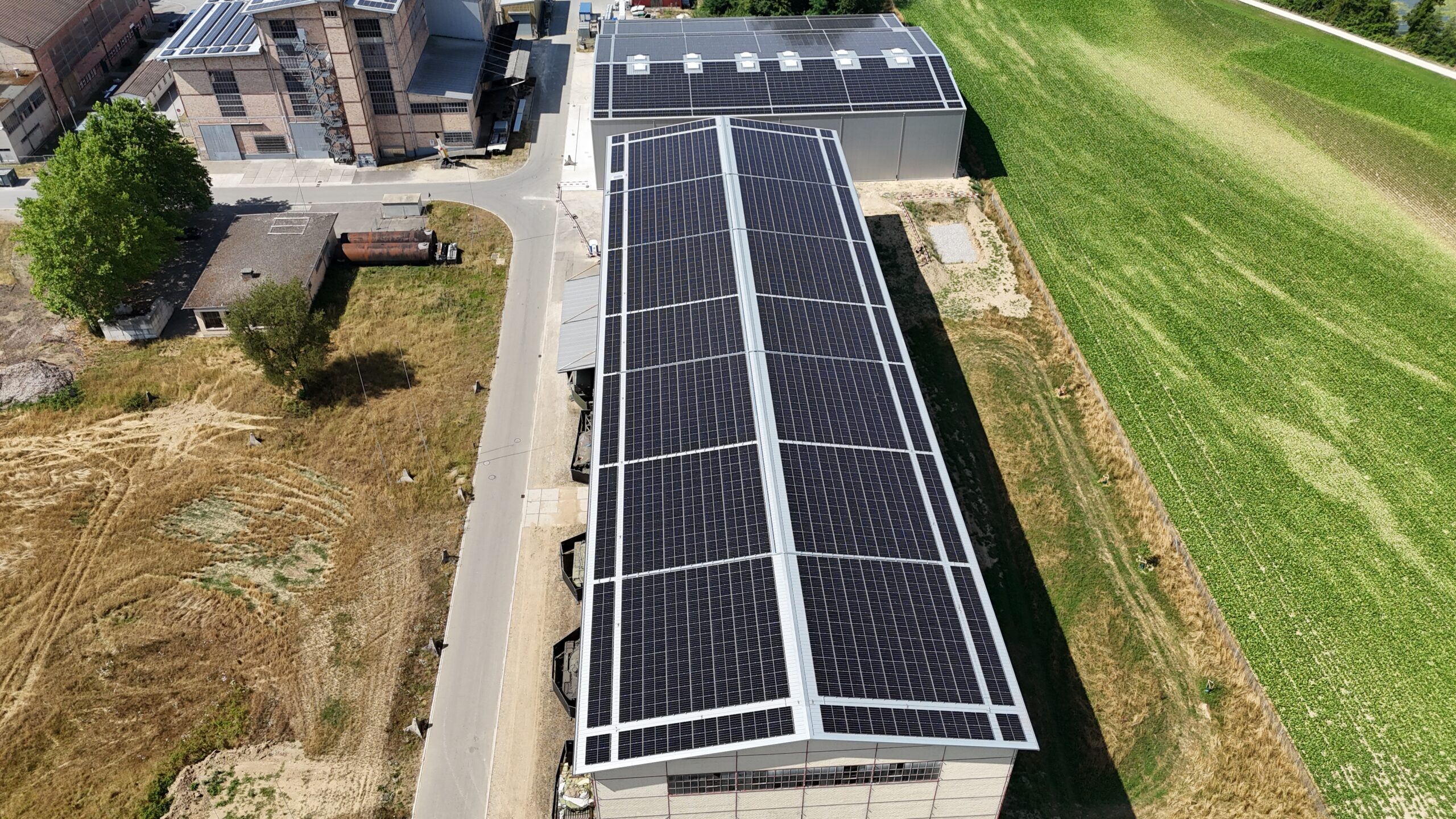 Aerial view of a large solar panel array installed on the roof of an industrial building, with a road and green field nearby.