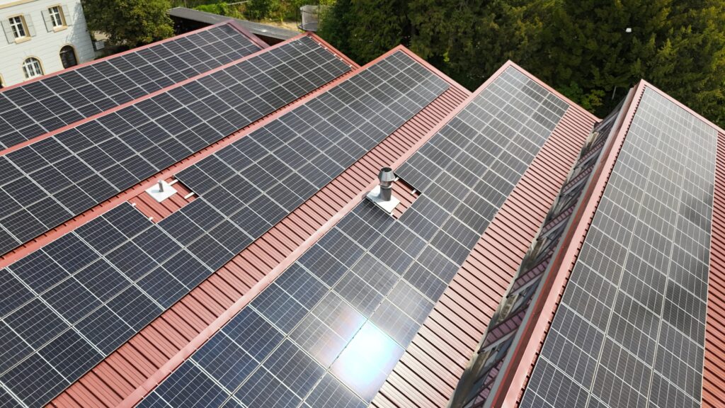 Aerial view of stacked dark solar panels on a red sloped roof with green trees in the background.