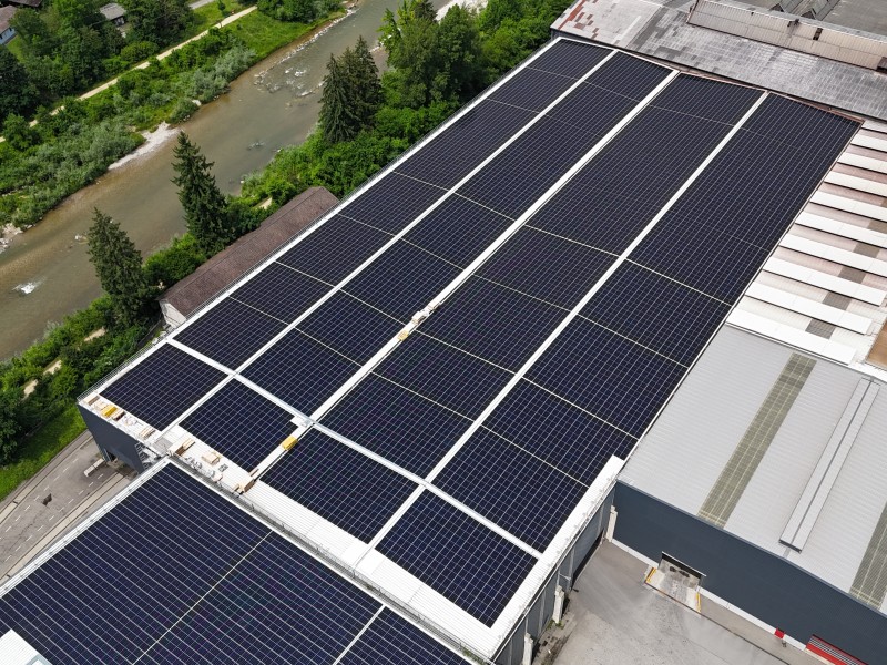 Aerial view of a flat rooftop covered with dark solar panels arranged in rows, with a river and trees nearby.