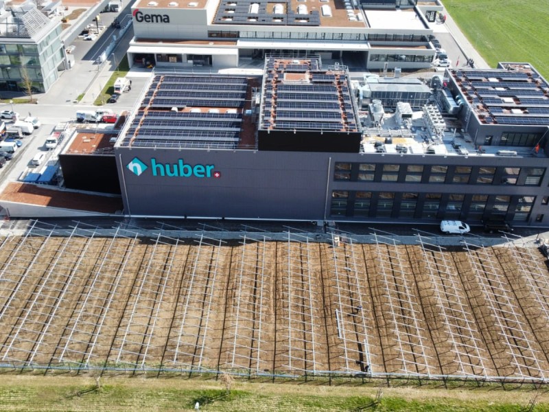 Aerial view of a modern industrial building with the 'Huber' logo, solar panels on the roofs, and an adjacent field with irrigation lines visible in the foreground.