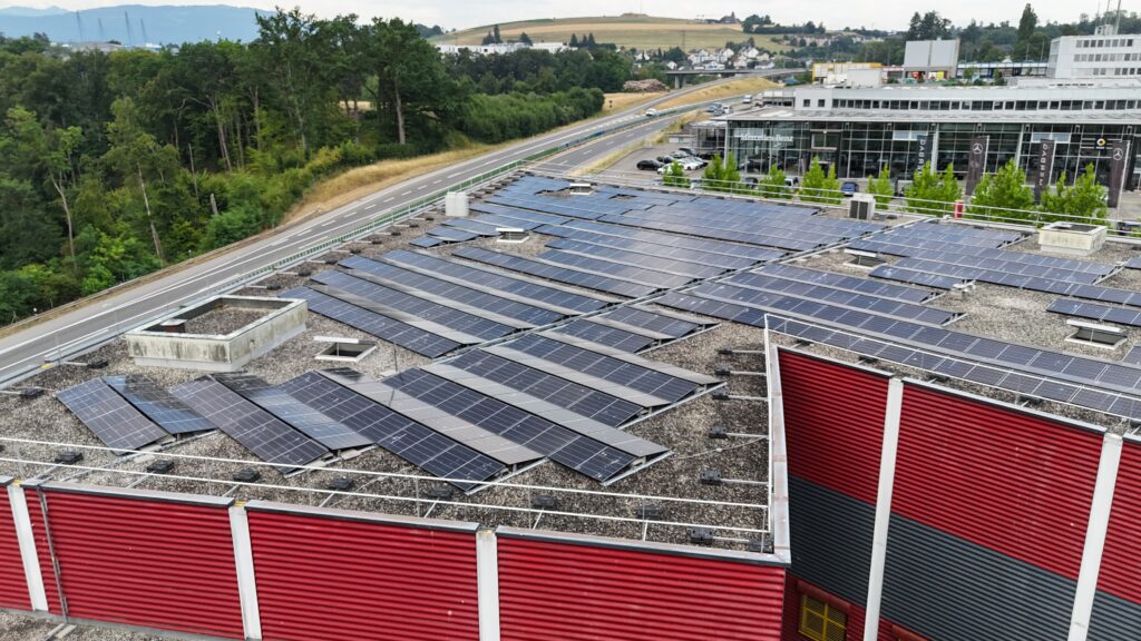 Rooftop covered with rows of solar panels on a modern commercial building, with a highway and green trees in the background.