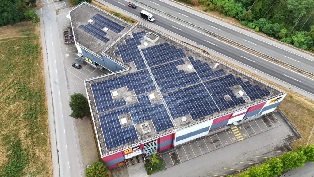 Aerial view of a commercial building with extensive solar panels on the roof, next to a highway and a parking lot.