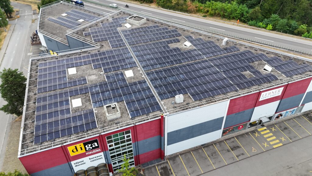 Aerial view of a commercial roof heavily covered with solar panels and a nearby parking lot adjacent to a road.