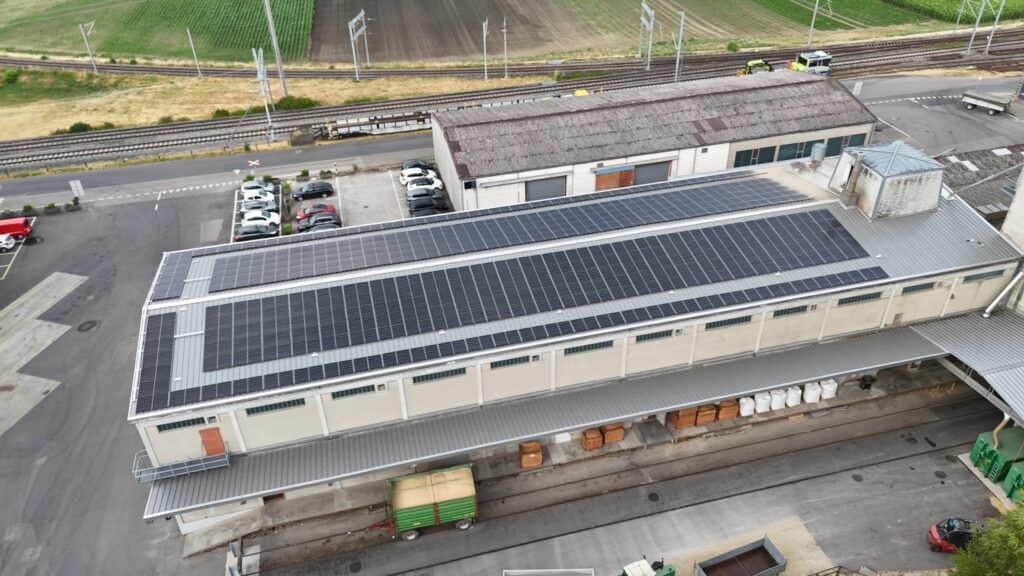 Aerial view of a factory with a large solar PV array on the roof, adjacent parking lot, and railway tracks in the background.