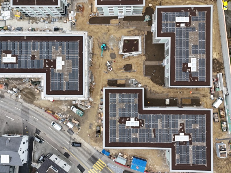 Aerial view of a construction site with L-shaped buildings, many solar panels on roofs, dirt yards, and construction equipment in the centerartifacts at street level.
