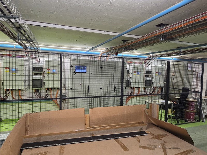 Electrical switchgear and control panels behind a safety mesh fence in an industrial room, with blue overhead pipes and tangled cables; cardboard box in the foreground.