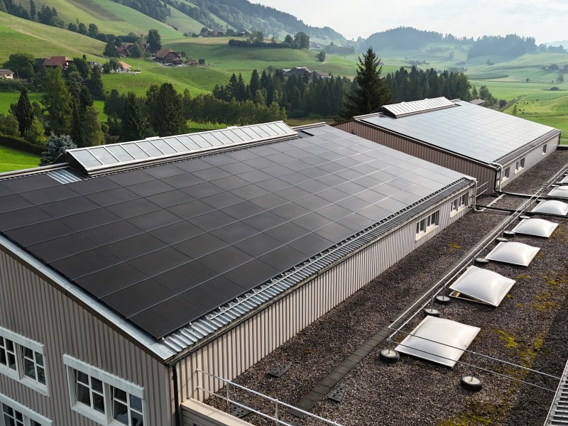 Solar panels covering flat rooftops of an industrial building with a green valley in the background fore‑grounded by hills and trees.