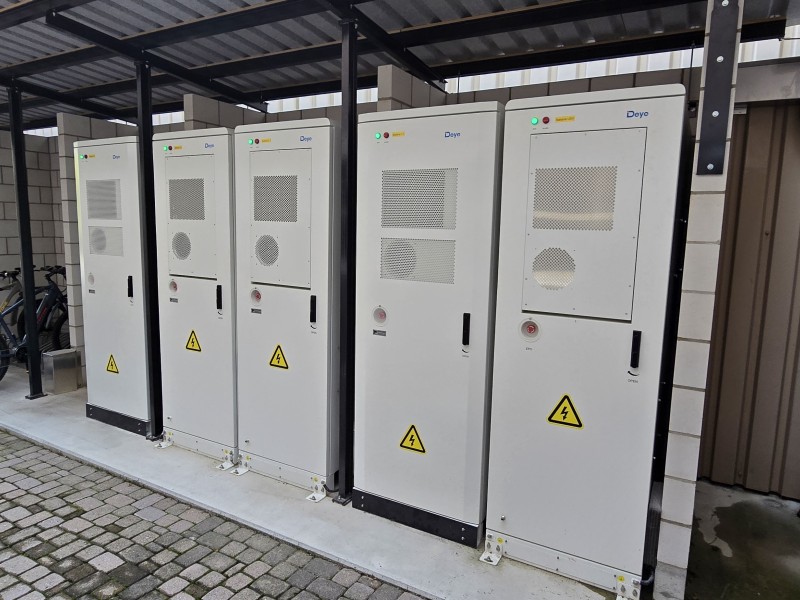 Row of gray outdoor electrical cabinets with high-voltage warning triangles under a metal canopy out front of a building.