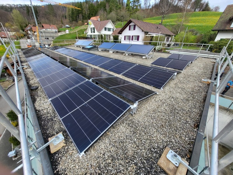 Rooftop solar panels installed on a gravel roof with rows of dark panels and surrounding houses in a green hillside neighborhood.