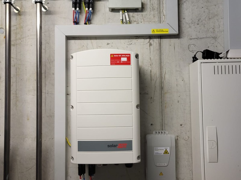 White solar inverter mounted on a concrete wall in a utility room, with cables above and an electrical cabinet nearby and warning labels visible.