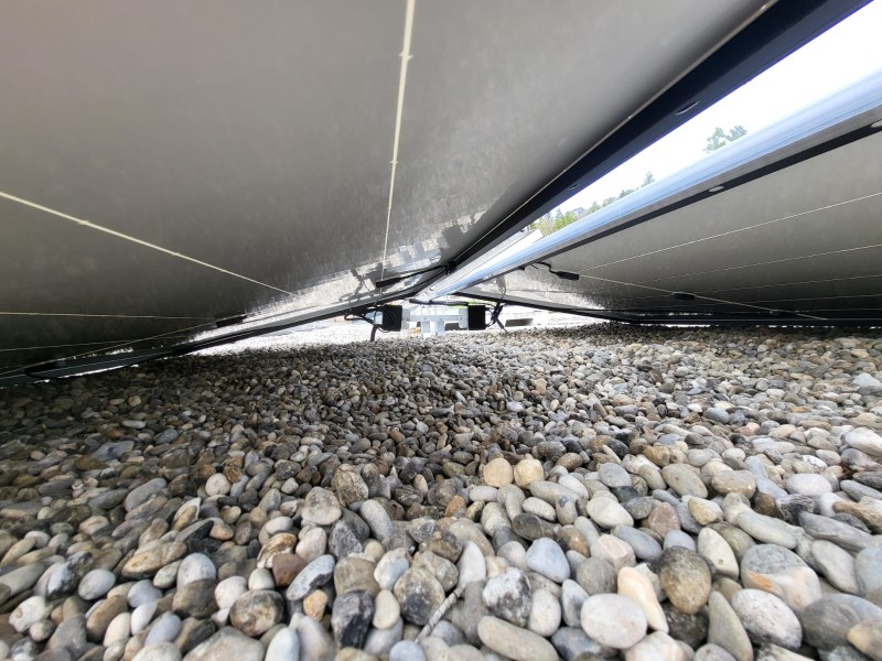 Ground-level view under a row of solar panels resting on a gravel surface, showing their metal framing and wiring.