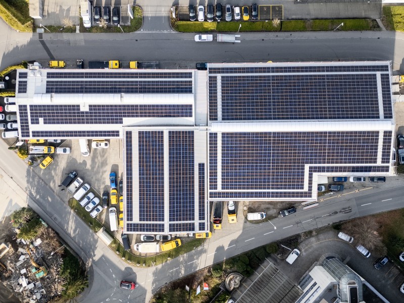 Aerial view of a large commercial building with extensive rooftop solar panels and surrounding parking lots and streets nearby.