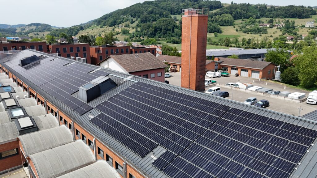 Factory rooftop covered with dark solar panels, brick buildings, and a tall brick chimney with a hilly, green landscape in the background