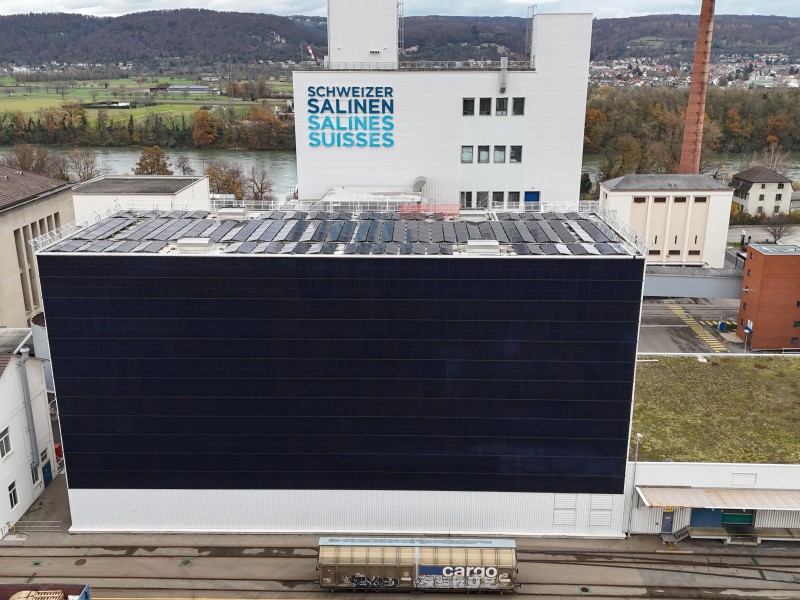 Large industrial complex with a dark solar-panel-clad building in the foreground, a white building behind displaying 'Schweizer Salinen / Salines Suiisses', and a cargo train on tracks in the foreground; hills and river in the distance.