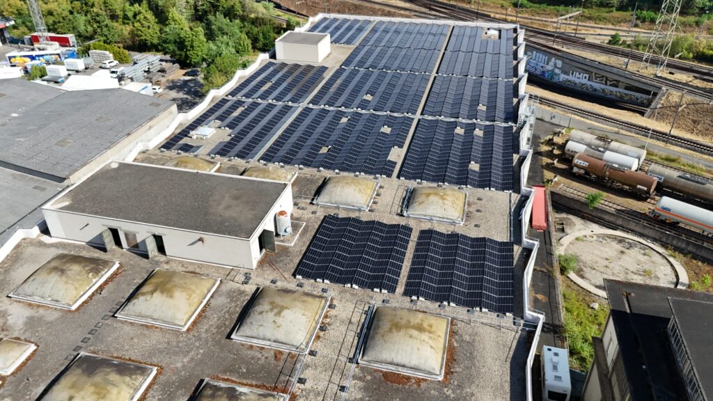 Aerial view of a large industrial rooftop covered with solar panels, with rail tracks and train cars beside.