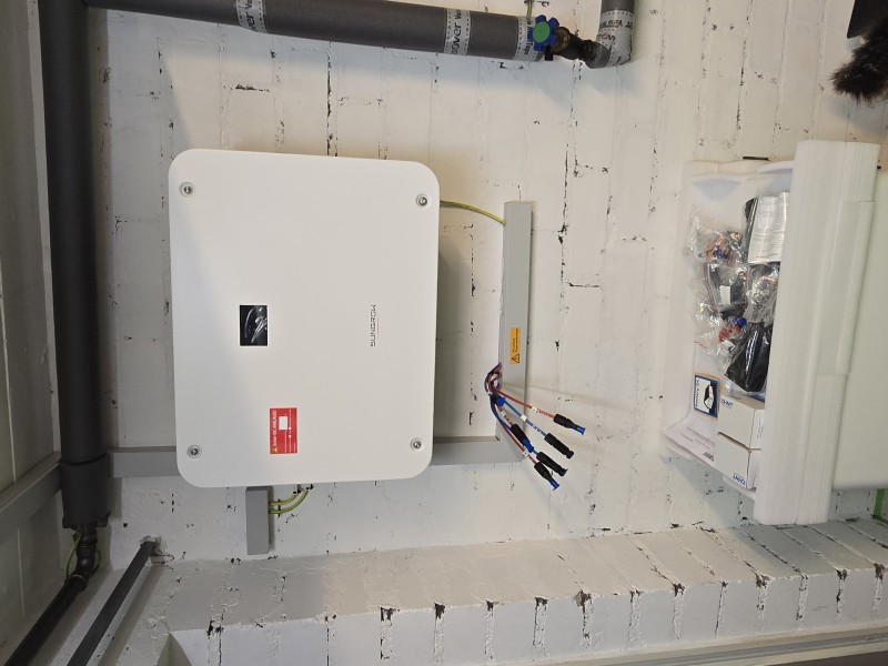 White electrical inverter mounted on a white brick wall with several color-coded cables and connectors coming out of a corded bundle nearby.