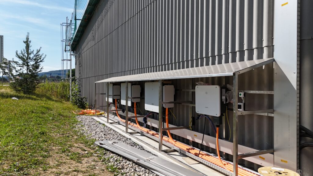 Electrical equipment and orange cables mounted on a metal-framed platform beside a corrugated gray building, outdoors on gravel ground far from vegetation.