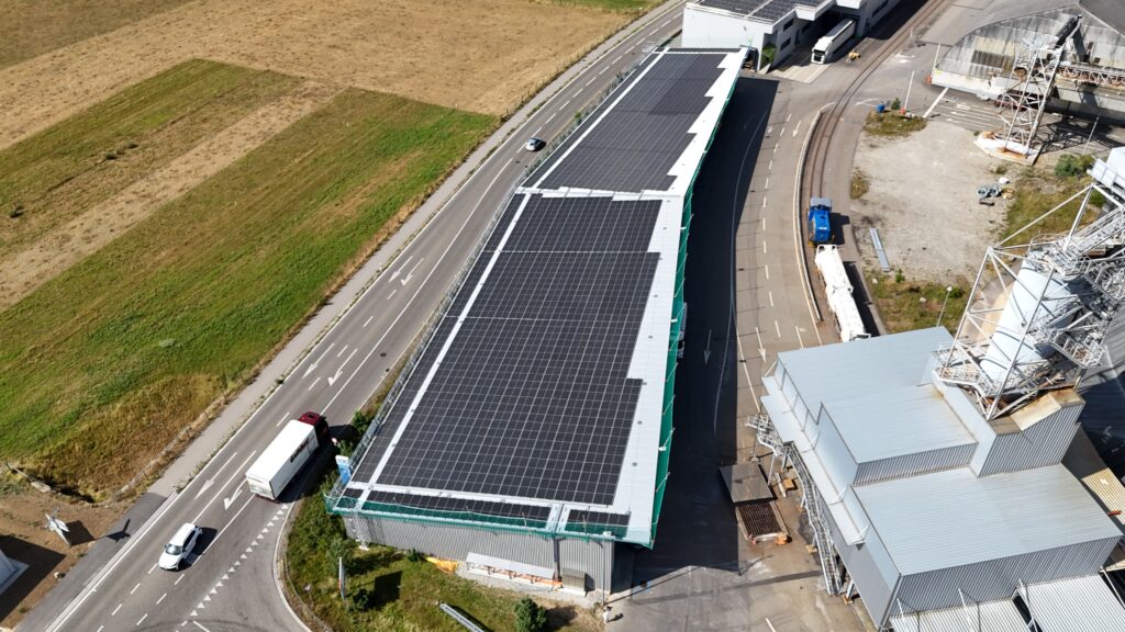 Aerial view of an industrial building with a solar-panel roof, next to roads and a loading=
