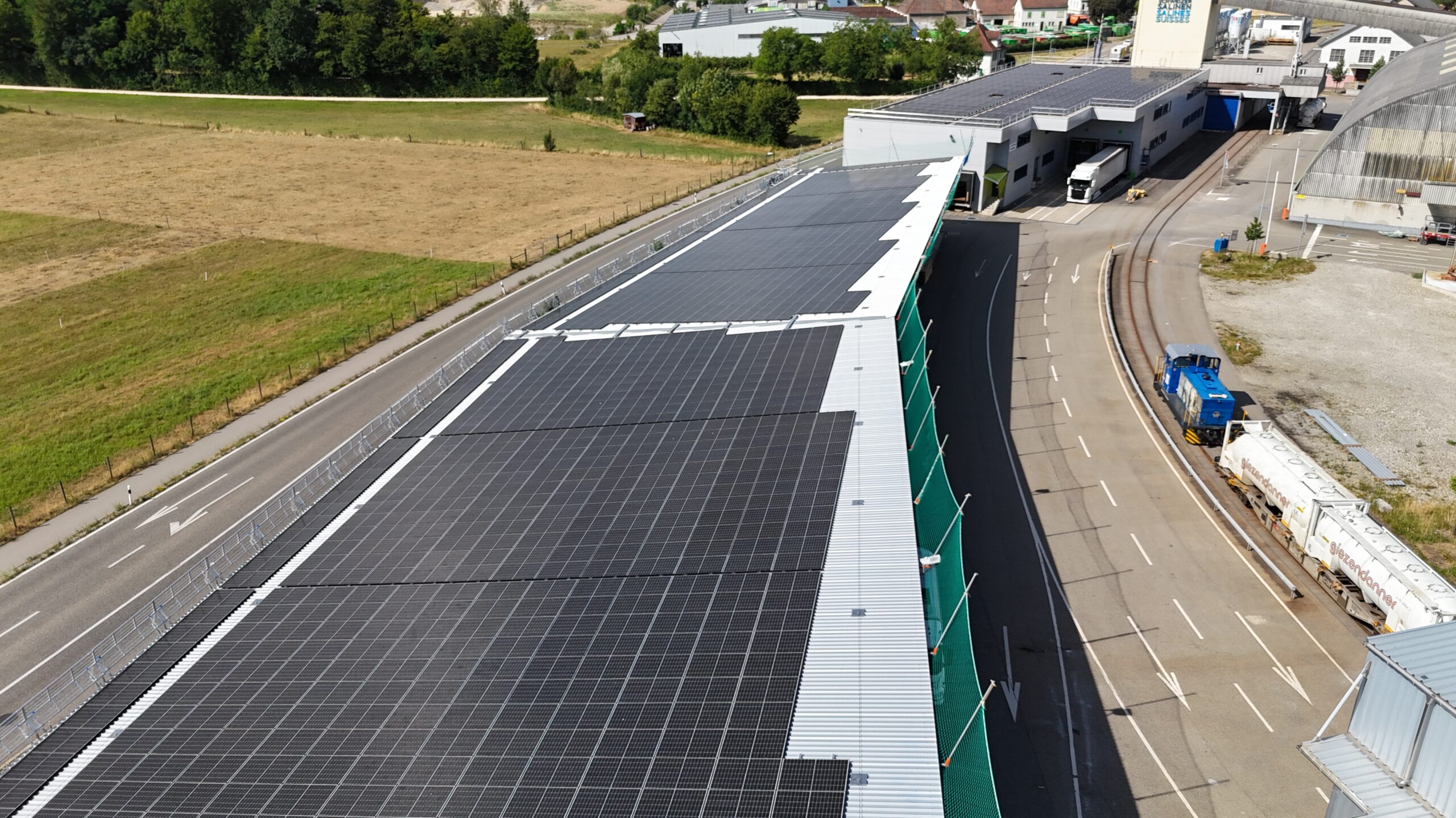 Aerial view of a solar-panel-covered roof running alongside a road, with trucks and industrial buildings nearby.