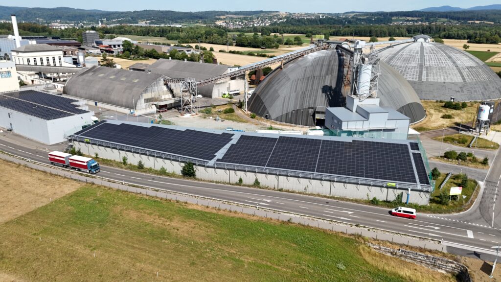 Aerial view of an industrial site with large curved-roof buildings, solar panels on rooftops, and trucks on a nearby road