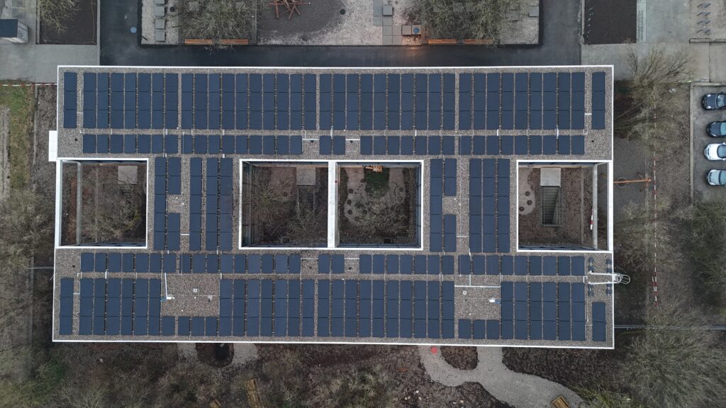 Aerial view of a large flat roof fully covered with dark solar panels arranged in neat rows around a central courtyard area inside a U-shaped building.
