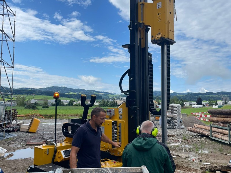 Yellow drilling rig at a construction site with two workers operating controls and wearing safety gear against a rural landscape backdrop