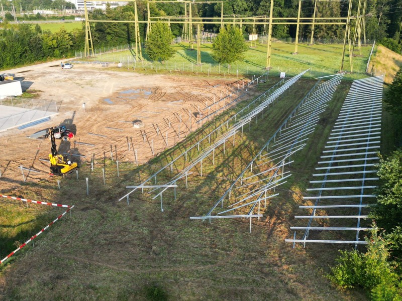 Outdoor sports venue under construction with metal bleacher framework and an excavator on a dirt field nearby, fenced area nearby.