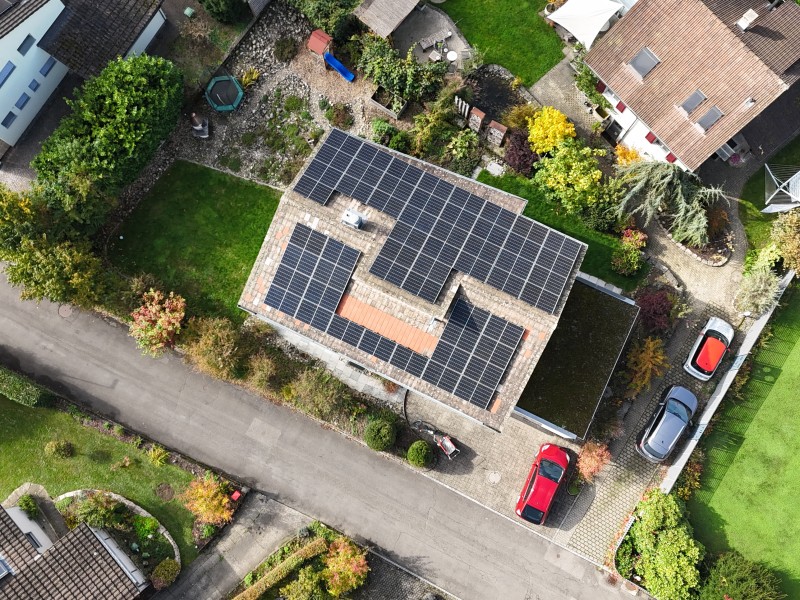 Aerial view of a residential property with solar panels covering the flat roof, cars parked in the driveway, and well-kept surrounding gardens and neighboring homes.