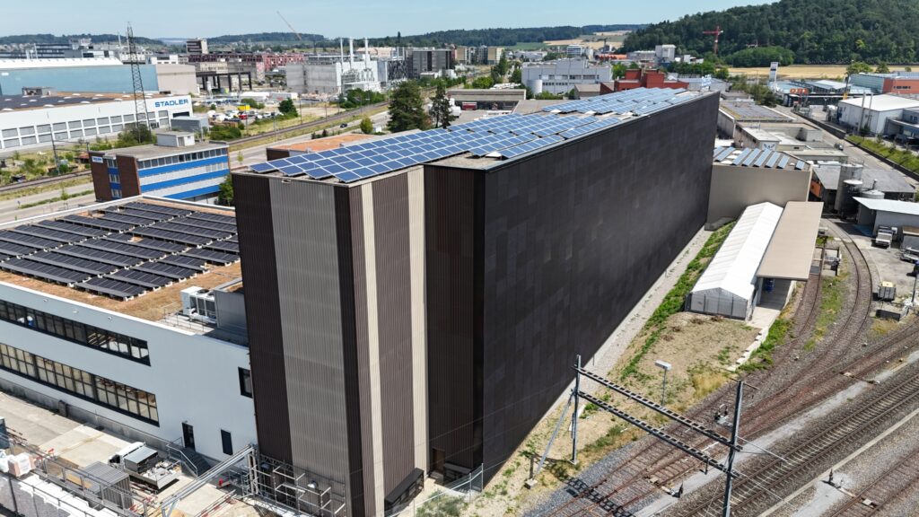 Rooftop solar panels array on a tall dark building beside railway tracks in an urban industrial area.